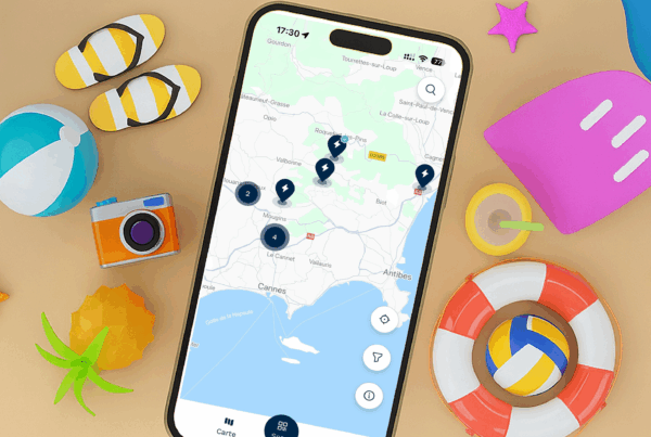 Un smartphone affichant l'app DRIVECO et une carte des stations de recharge autour de la région de Nice, dans un décors de vacances.