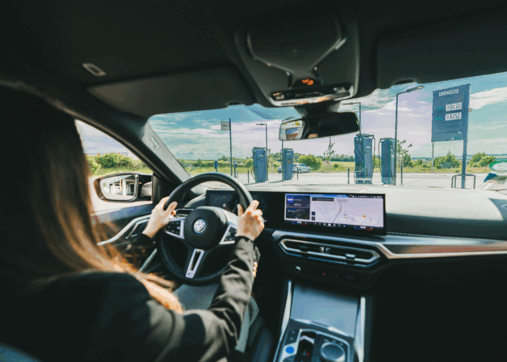 Une femme au volant d'une BMW i4 100% électrique, devant une station de recharge électrique DRIVECO.
