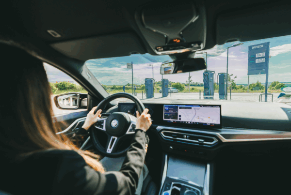 Une femme au volant d'une BMW i4 100% électrique, devant une station de recharge électrique DRIVECO.
