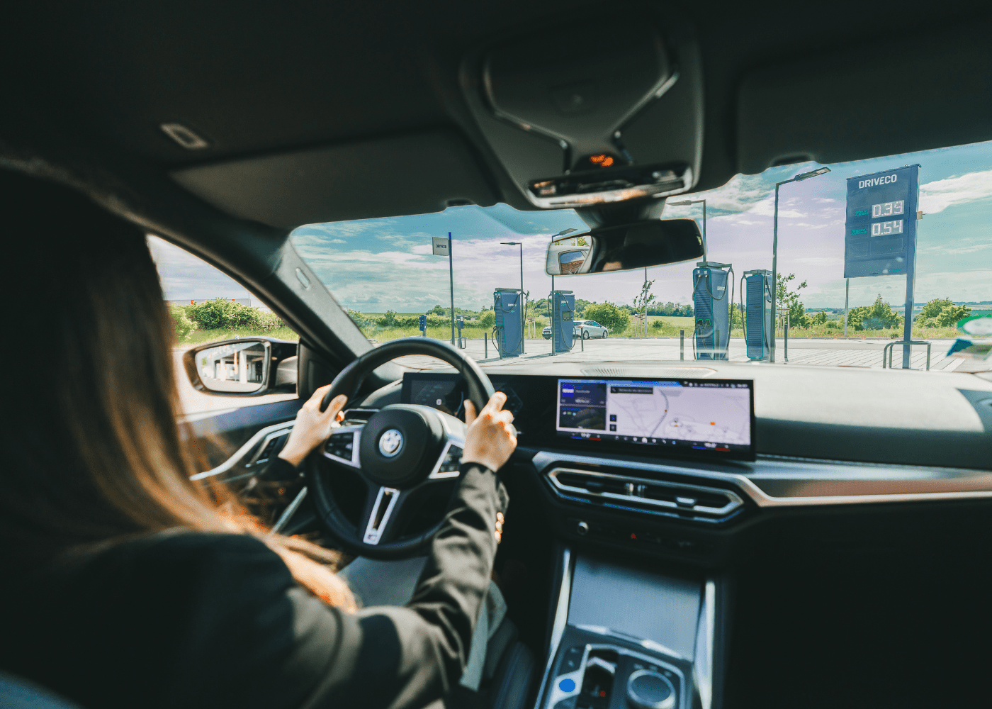 Une femme au volant d'une BMW i4 100% électrique, devant une station de recharge électrique DRIVECO.