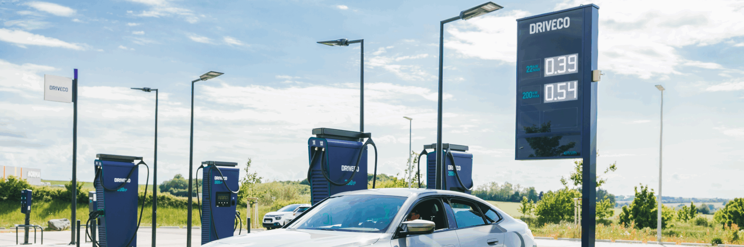 Voiture électrique en charge sur une station DRIVECO moderne, équipée de bornes rapides et d’un totem d’affichage des tarifs sous un ciel ensoleillé.