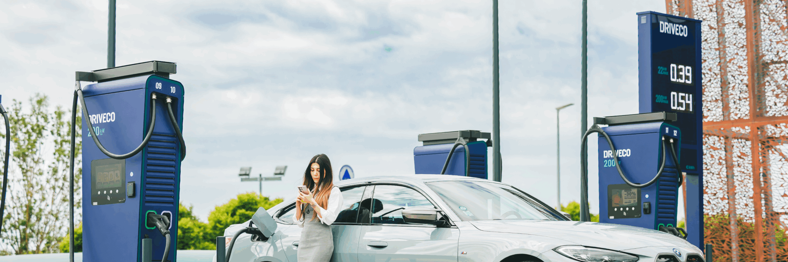 Une femme se tient devant sa voiture électrique stationnée et en charge sur une borne DRIVECO 200 kW.