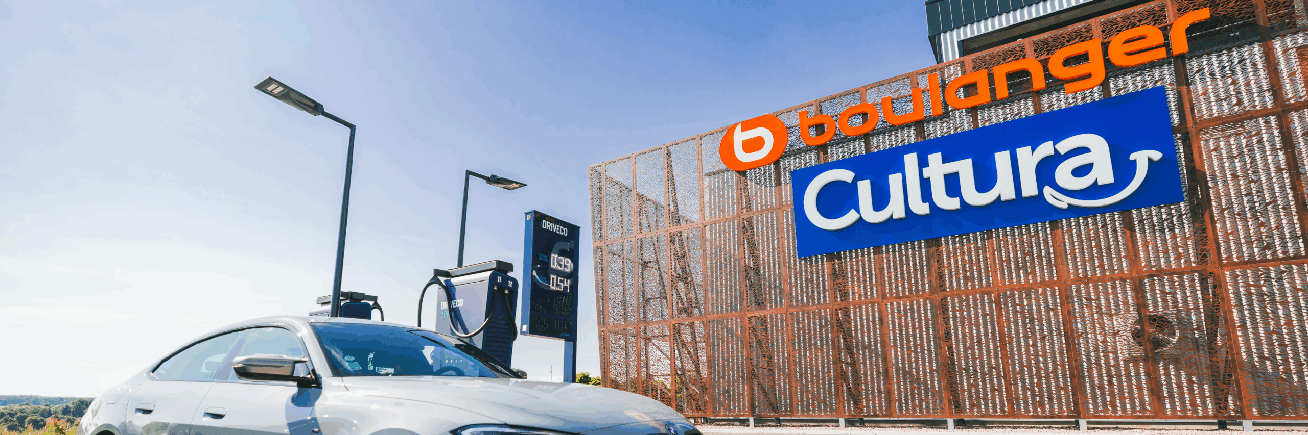 Borne de recharge Driveco installée sur le parking d’un centre commercial, avec une voiture électrique en cours de charge devant les enseignes Boulanger et Cultura.