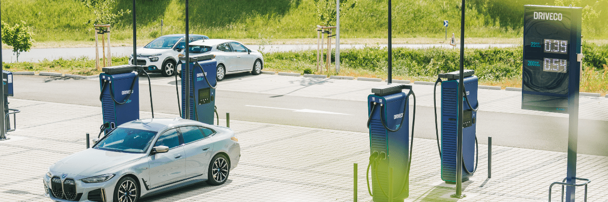 Vue d’ensemble d’une station de recharge DRIVECO équipée de plusieurs bornes rapides, avec une BMW électrique grise branchée au premier plan et des voitures garées en arrière-plan.