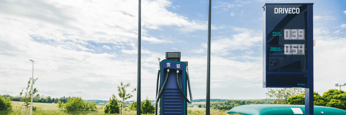 Borne de recharge DRIVECO installée en extérieur, équipée de deux câbles, avec à droite un totem affichant les tarifs de recharge en kWh.