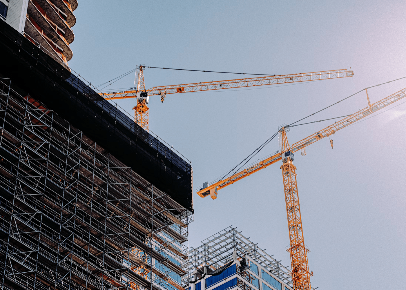 Deux grues de chantier jaunes surplombant un immeuble en construction entouré d’échafaudages métalliques, avec un ciel bleu dégagé en arrière-plan.