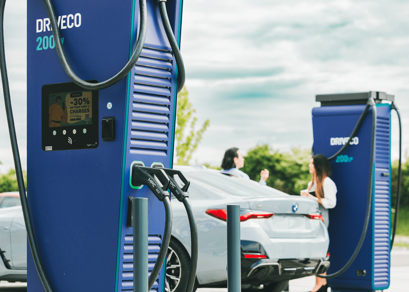 Bornes de recharge rapides DRIVECO 200 kW en fonctionnement, avec un véhicule électrique branché et deux personnes discutant à proximité sur un parking en plein air.