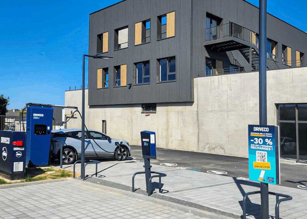 Station de recharge DRIVECO avec bornes rapides 200 kW et bornes 22 kW, située devant un bâtiment moderne. Une voiture électrique grise est en cours de recharge. Panneau promotionnel DRIVECO offrant -30 % sur les trois premières recharges.