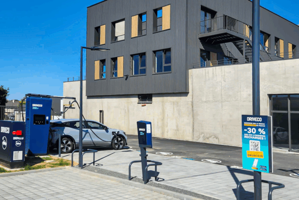 Station de recharge DRIVECO avec bornes rapides 200 kW et bornes 22 kW, située devant un bâtiment moderne. Une voiture électrique grise est en cours de recharge. Panneau promotionnel DRIVECO offrant -30 % sur les trois premières recharges.