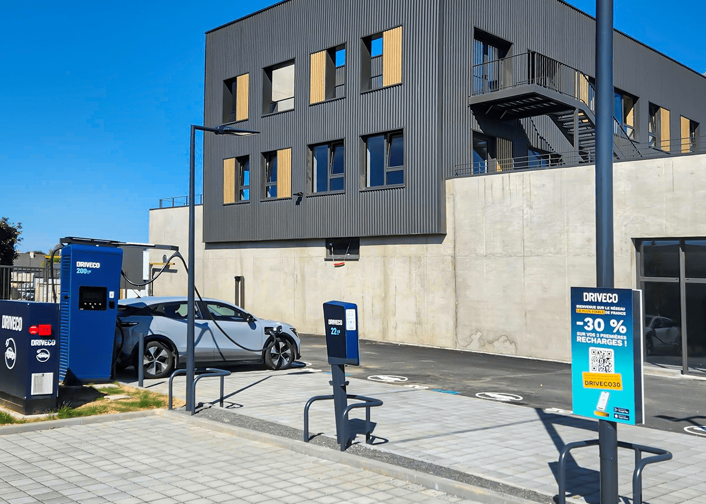 Station de recharge DRIVECO avec bornes rapides 200 kW et bornes 22 kW, située devant un bâtiment moderne. Une voiture électrique grise est en cours de recharge. Panneau promotionnel DRIVECO offrant -30 % sur les trois premières recharges.