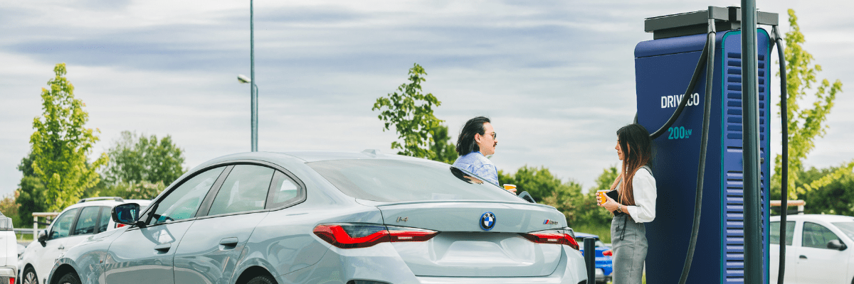 Deux personnes discutent près d’une voiture électrique BMW en charge sur une borne rapide DRIVECO de 200 kW, sur un parking arboré par temps couvert.