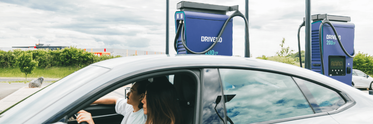 Conductrice et un passager dans une voiture électrique blanche stationnée à une station de recharge rapide DRIVECO avec bornes de 200 kW en arrière-plan.