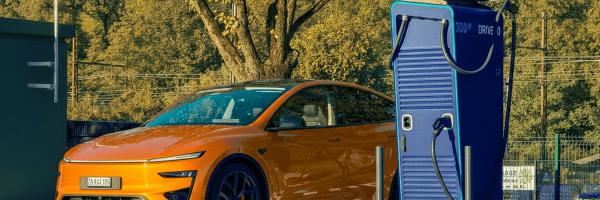 Voiture électrique orange en train de se recharger sur une borne de recharge rapide DRIVECO, dans un parking entouré d’arbres en automne.