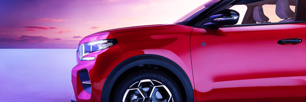 Vue rapprochée de l’avant et du côté d’un Citroën e-C3 SUV rouge électrique, éclairé par une lumière douce au coucher du soleil, mettant en valeur les lignes modernes du véhicule et son phare avant.