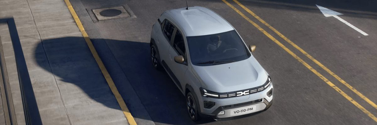 Vue aérienne d’une Dacia Spring blanche roulant sur une route urbaine, entre deux lignes jaunes, sous un soleil créant une ombre nette du véhicule sur le trottoir.