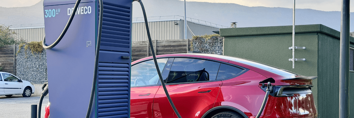 Voiture électrique rouge branchée à une borne de recharge rapide 300 kW DRIVECO, vue de profil, sur un parking extérieur avec montagnes en arrière-plan.