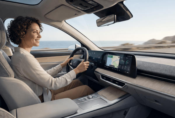 Femme souriante conduisant une voiture moderne avec tableau de bord numérique sur une route côtière au bord de l'océan.