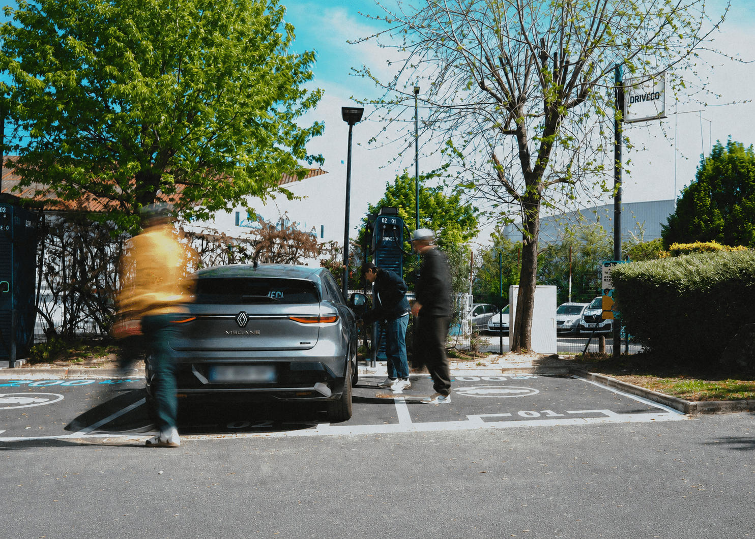 Une voiture électrique garée devant une borne DRIVECO, avec des personnes passant autour du véhicule
